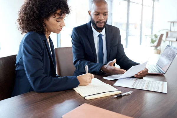 an image showing a female and a male sitting side by side, with papers on the table, they're looking the papers on a table and talking about them and taking notes.